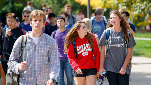 Two female students talk and laugh while walking along a crowded university path.