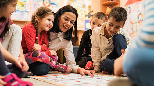 A female adult interacts with a group of children on the floor of a classroom.