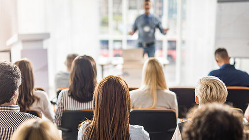A man talks to a group of adults in a bright room.